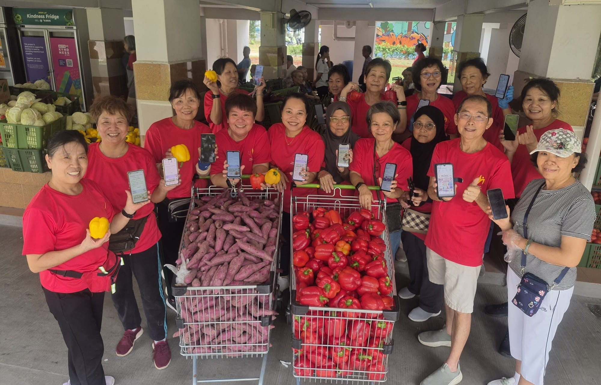 A group of Neighbourhood Kakis volunteers in red t-shirts pose cheerfully with trolleys filled with purple sweet potatoes and red capsicums, holding up their smartphones at a void deck.